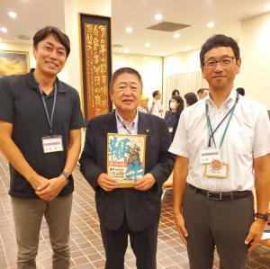 市長が「鞭牛 拓け命の道を」のパンフレットを持ち、両脇に男性2名が立って撮影した写真