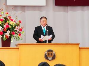市長が花が飾られたステージ上の演台で、用紙を広げて式辞を述べている写真