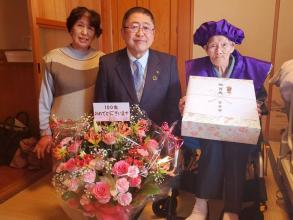 誕生日を祝う花かごを前にして、紫色のちゃんちゃんこと頭巾姿の中嶋磨知さんがお祝いの品物を持ち、娘さんと市長の3人で撮影した写真