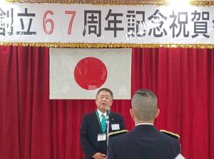 赤い幕に日本国旗が吊るされた会場で、マイクに向かい話をする様子の市長とそれを聞く自衛隊員の写真
