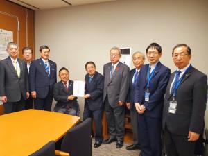 議員会館事務所の室内で椅子に座っている男性と市長が一緒に陳情書を持って中央に立ち、7名の関係者が横一列に並んでいる写真