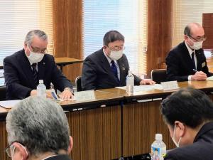 向かい合わせに置かれた茶色いテーブルと椅子に座り、机の上に置かれた資料を見る様子の市長と関係者の男性が写っている写真