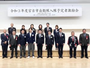「令和3年度宮古市自衛隊入隊予定者激励会」と書かれた看板が後方の壁に吊るされた会場で、前列に赤い胸章を着けた市長を含めた9名が立ち、1段上がった後列に6名が立って並んでいる記念写真