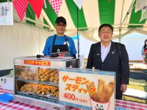 白と緑色のストライプ柄のテントの中に「サーモンスティック400円」と書かれたポップと商品が陳列され、古須賀商店の店主の方と市長が並んでいる写真