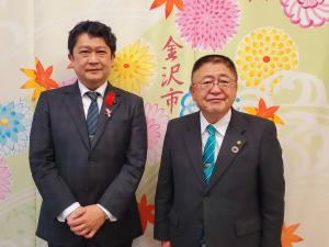 両手を身体の前で組んでいる石川県金沢市の村山卓市長と山本市長が横並びで写る記念写真
