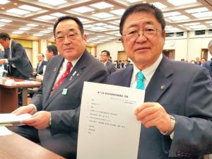 防災対策特別委員会の席で隣席の関係者の男性と、左手で委員会の資料を持ちカメラに見せている市長の写真