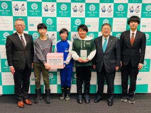 宮古市のバックパネルをバックに宮古小学校の児童3名が能登半島地震災害義援金の箱を持って市長と関係者の方にはさまれて記念撮影をしている写真