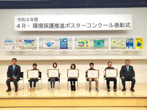 令和4年度4R環境保護推進ポスターコンクール表彰式と書かれた横断幕の下に入賞したポスターが飾られ、それを背景に表彰された児童5人がそれぞれ表彰状を持って座り、市長と副市長の 間に並んで撮影した写真