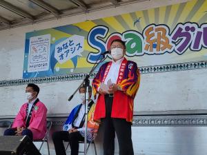 法被を着てスタンドマイクの前で話をする様子の市長と、市長の斜め後ろで椅子に座っている法被を着た男性2人の写真