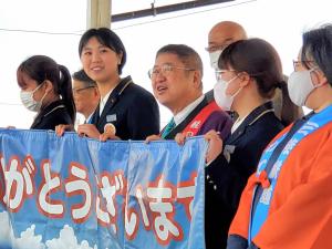 市長と女性3名が横断幕を持ち、横にオレンジ色の法被を着た女性が立ち、その後ろに関係者の方々が立っている写真