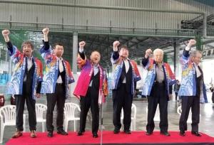 赤い絨毯が敷かれた台の上に立ち、右手を上にあげている様子の市長と5名の関係者の男性たちの写真