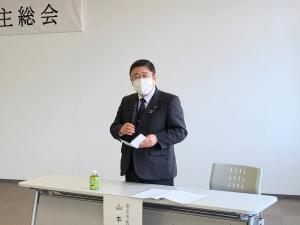 名前札が貼られたテーブルと椅子の間に立ち、右手に資料、左手にマイクを持ってマスクを着けた市長が話をしている様子の写真