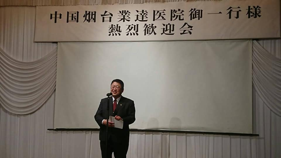 「中国烟台業達医院御一行様 熱烈歓迎会」と書かれた横断幕やスクリーンが設置された会場の中央で、市長が挨拶を行っている写真