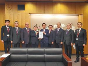 3人掛けのソファが設置された室内で総務大臣政務官の船橋利実氏と市長が一緒に要望書を持って中央に立ち、7名の関係者が横一列に並んでいる写真