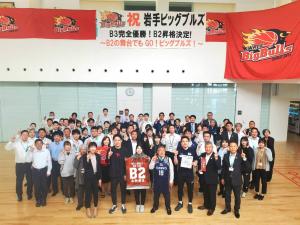 「祝 岩手ビッグブルズ」と書かれた横断幕の下で大勢の方たちが右手の人差し指を上に向けポーズを決めて写っている記念写真