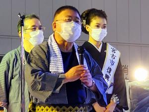 法被を着てハチマキを首にかけマイクを持った市長と、その後方に立つ男性と着物を着た女性を写した写真