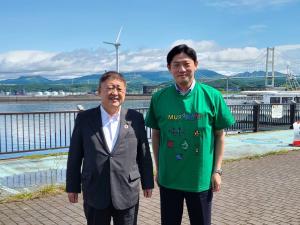 海を背にスーツを着た市長と緑色のTシャツを着た男性が並んで写っている写真