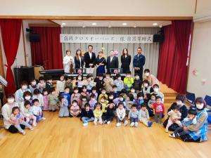 小さなステージに「盛岡クロスオーバーズ様 楽器寄贈式」と書かれた横断幕が掲げられ、寄贈された楽器の後ろに盛岡クロスオーバーズとグラッツィアのメンバー、関係者の男性と市長が立ち、手前の広場に田老保育所の先生や子供たちが座りっている写真