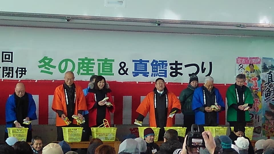 「第2回宮古・下閉伊冬の産直&真鱈まつり」と書かれた横幕と紅白幕が壁に貼られた会場前方に赤や青色などのカラフルな色の法被を着た関係者が立ち、両手に商品を持ったオレンジ色の法被を着た市長が大勢のお客さん達に向かって話をしているイベントの様子の写真