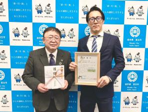 バックパネルの前で目録を持つ市長と、額縁を持つ眼鏡をかけスーツ姿の男性を写した写真