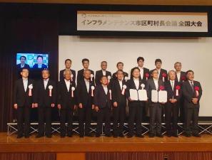 壇上の上でスーツを着て胸に胸章を付け2列に並んで記念撮影をする男性たちの写真