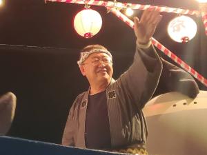明かりがついた提灯が掛けられた櫓の上で法被を着て頭にハチマキを巻き左手を上げ手を振る市長の写真