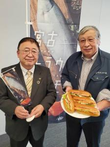 「宮古トラウトサーモン」の文字とサーモンの写真が載った幟の前で、小さな幟を両手で持つ市長とサーモンを使用したホットドッグが乗ったお皿を持つ男性が並んで立っている写真