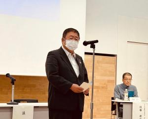 後方に大きなスクリーンが設置された定期総会の会場で、マイクスタンドの前に立ち話をしている市長の写真