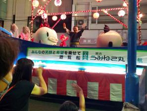 両手を上に掲げる市長と、サーモンくんとみやこちゃんが櫓の上立ち、それを観客が写真に撮っている様子の写真