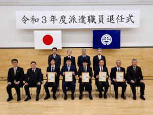 「令和3年度派遣職員退任式」と書かれた横断幕と、国旗、宮古市の市町村旗が吊り下げられた会場で、左から男性が1名、市長、額縁を持った5名と1名の男性が前列で椅子に座り、後列に4名の男性が立って横に並んでいる写真
