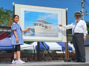 ボードに貼り付けられた遊覧船の写真を、ボードの両側からそれぞれ指さす女の子と制帽を被った市長の写真