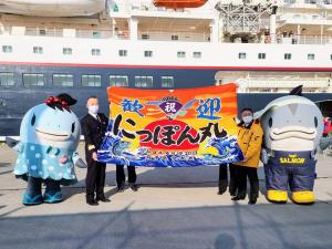 宮古港に入港した客船「にっぽん丸」をバックに、船長と市長が「歓迎にっぽん丸」と書かれ荒れ狂う波と日の出の様子が描かれた大きな旗を持ち、両端に宮古市のマスコットキャラクターサーモンくんとみやこちゃんが立ち並んでいる写真