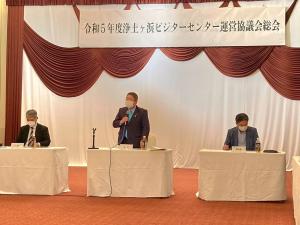 「令和5年度浄土ヶ浜ビジターセンター運営協議会総会」と書かれた横断幕の下で、白いテーブルクロスが引かれた机の後ろに立ちマイクを持って話す男性と、その両側で椅子に座っている男性2名の写真