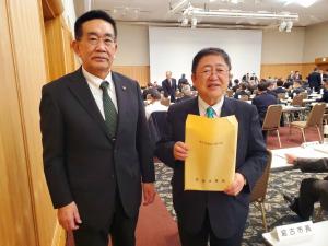 前方に大きなスクリーンが設置された「理事・評議員合同会議」の会場で、大きな封筒を持っている市長と関係者の男性が横一列に並んでいる写真