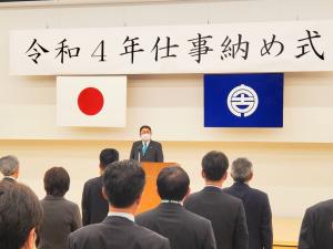 令和4年仕事納め式と書かれた横断幕の下で市長が演台で、立っている参加者の方々に式辞を述べている写真