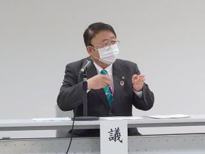 机に置かれたスタンドマイクの前で椅子に座り手を胸の前にあげて動かしながら話している様子の市長の写真