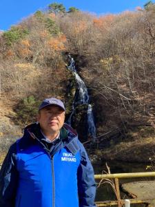 小高い山の中腹から水が流れている滝の前に市長が立っている写真
