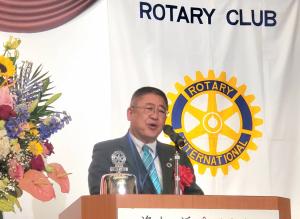 ロータリークラブのロゴマークがあしらわれた幕をバックに、檀上に立つ市長が挨拶を行っている様子をアップで撮影した写真