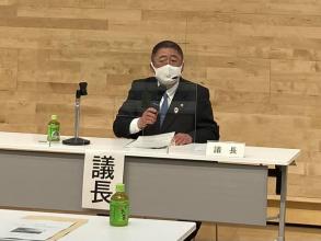 議長と書かれた紙が貼られた机で市長が総会で意見を述べている写真