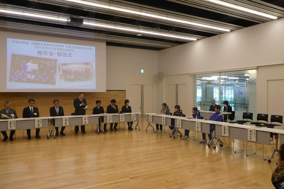 「報告会・解団式」と書かれた資料が映し出さされた大きなスクリーンが設置された室内に、長机をロの字型に設置された席に小中学生や関係者が座り、前方中央の席から立ち上がった市長が子供たちの方向を向いて話をしている様子の写真