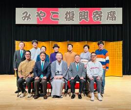 暗幕に「みやこ復興寄席」と書かれた横断幕と舞台に金屏風が飾られ関係者12名の方々が2列に並んで記念撮影をしている写真