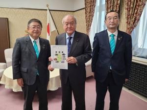 国旗が設置された室内で、財務省の鈴木俊一大臣と市長が一緒に要望書を持って立ち、右側に関係者の男性と横一列に並んでいる写真