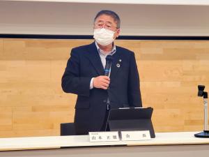 机に置かれたタブレットの前で立ち右手にマイクを持って話す市長の写真