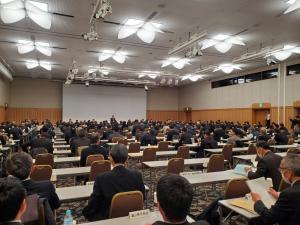 2名ずつかけられる長机が沢山並べられ、前方にはスクリーンが設置された会議室内を後方から写した写真