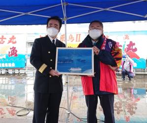 客船飛鳥2の船長と法被を着た市長が一緒に額縁に入った飛鳥2が航行している様子の写真を持ち横に並んでいる記念写真