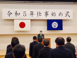 「令和5年仕事始め式」と書かれた横断幕の下で、演壇に立つ市長の話を聞いている様子を後方から撮影した写真