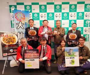 大仙市の旗が置かれた宮古市のバックボードの前で、関係者8名がポスターや商品を持って2列に並んでいる写真