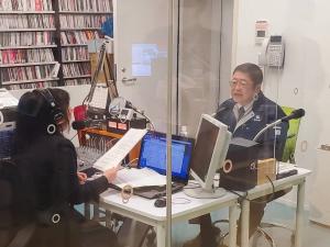 右手に資料を持ちヘッドホンを付けた女性の向かい側で、マイクに向かい話している様子の市長の写真