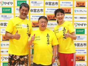 「宮古市」の文字や企業の広告が掲載されたバックパネルの前で、市長を中心に並ぶ高田延彦さん、向井亜紀さんがお揃いの黄色のTシャツを着て片手の親指を上に向けポーズをとっている記念写真