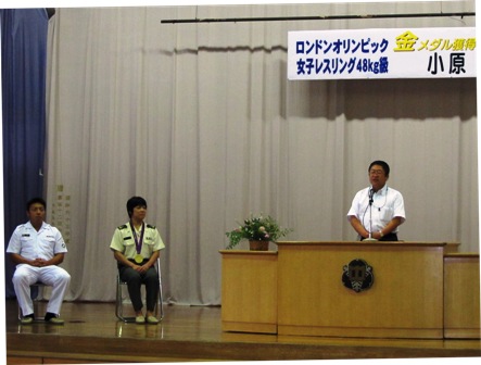 市長が演壇の前に立ちマイクを使い話しており、その横で上下白い制服をきた男性と上が薄い黄色、下が深緑色のズボンをはいた首からメダルをかけた女性がパイプ椅子に座っている写真